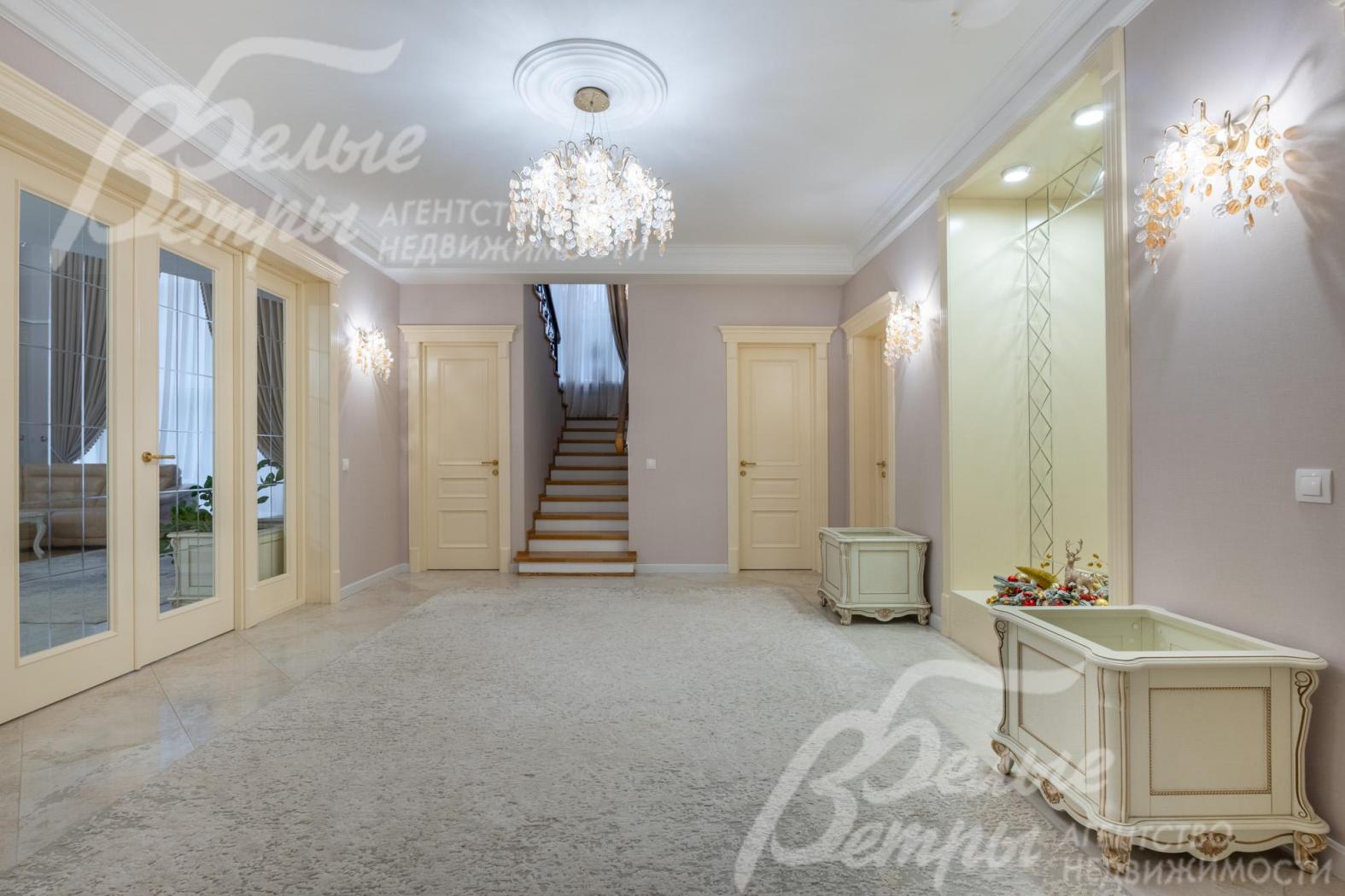 В просторном холле дома уложен уложен керамогранит (тёплый пол), ковёр, интерьер дополняют эксклюзивные скамьи-тумбы, большое зеркало и дорогостоящая люстра